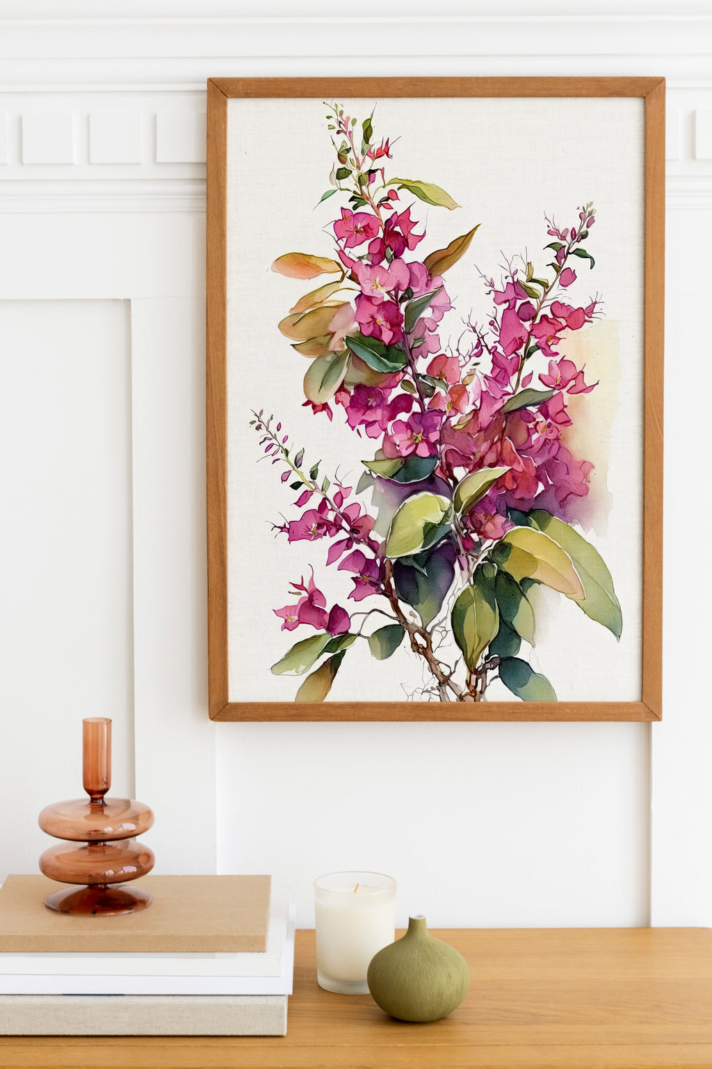 a framed painting of a vibrant floral arrangement hanging on a white wall above a wooden table. The table also holds a stack of books, a small green vase, and a white candle.