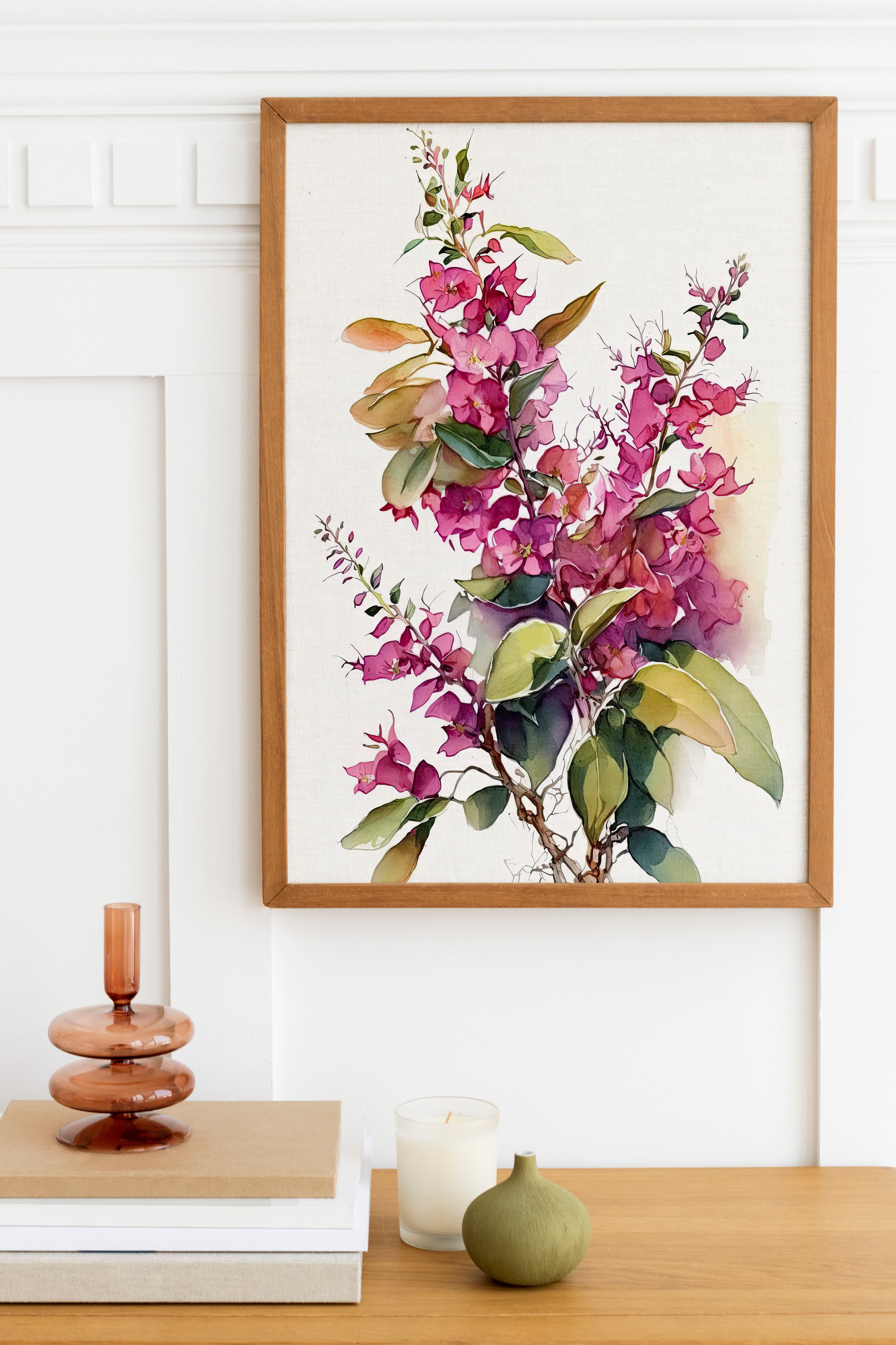 a framed painting of a vibrant floral arrangement hanging on a white wall above a wooden table. The table also holds a stack of books, a small green vase, and a white candle.