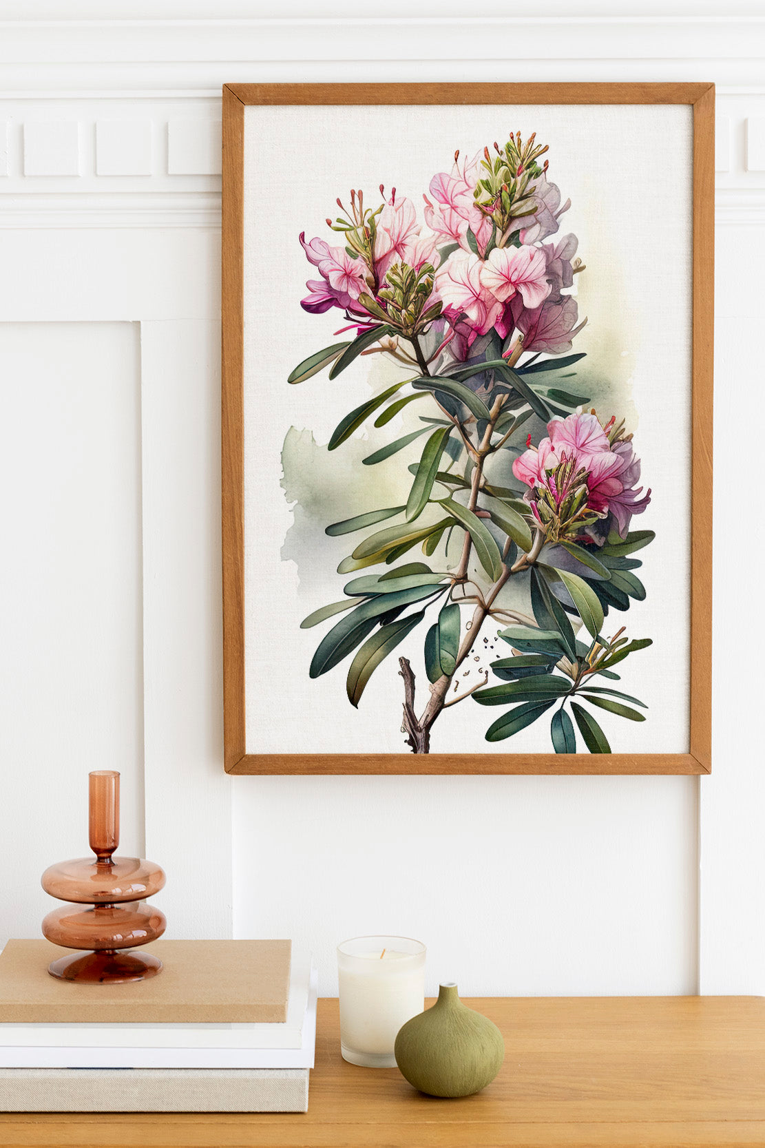 a framed painting of a pink flower on a white wall, with a wooden shelf below it holding a stack of books, a small green vase, and a white candle.