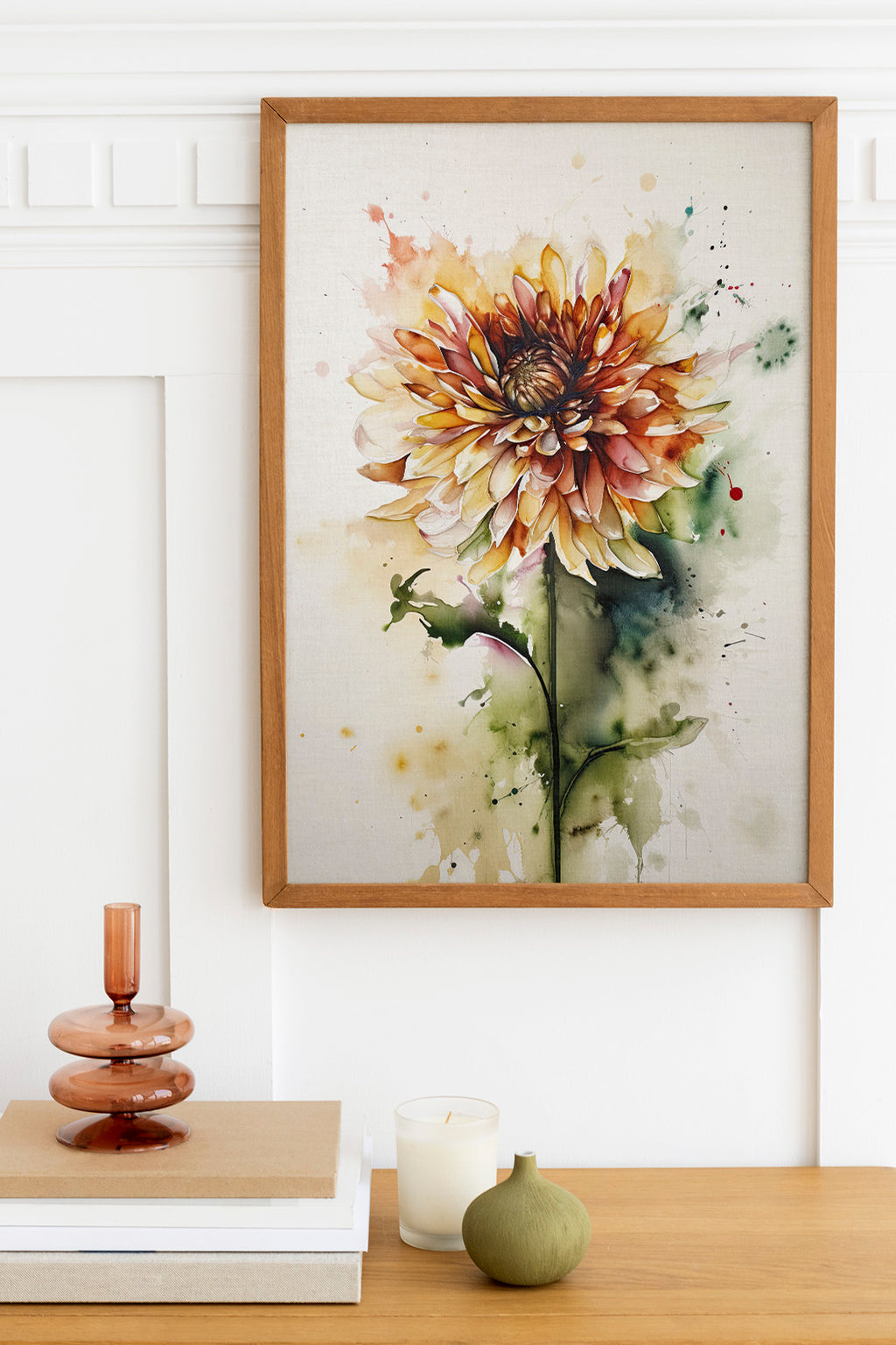 a framed watercolor painting of a vibrant yellow and orange flower, hanging on a white wall above a wooden table. The table also holds a stack of books, a small green vase, and a white candle.