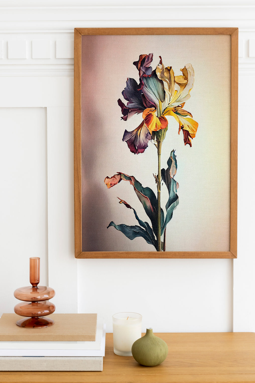a framed painting of a vibrant, colorful flower hanging on a wall above a wooden table. On the table, there is a stack of books, a small green vase, and a white candle.
