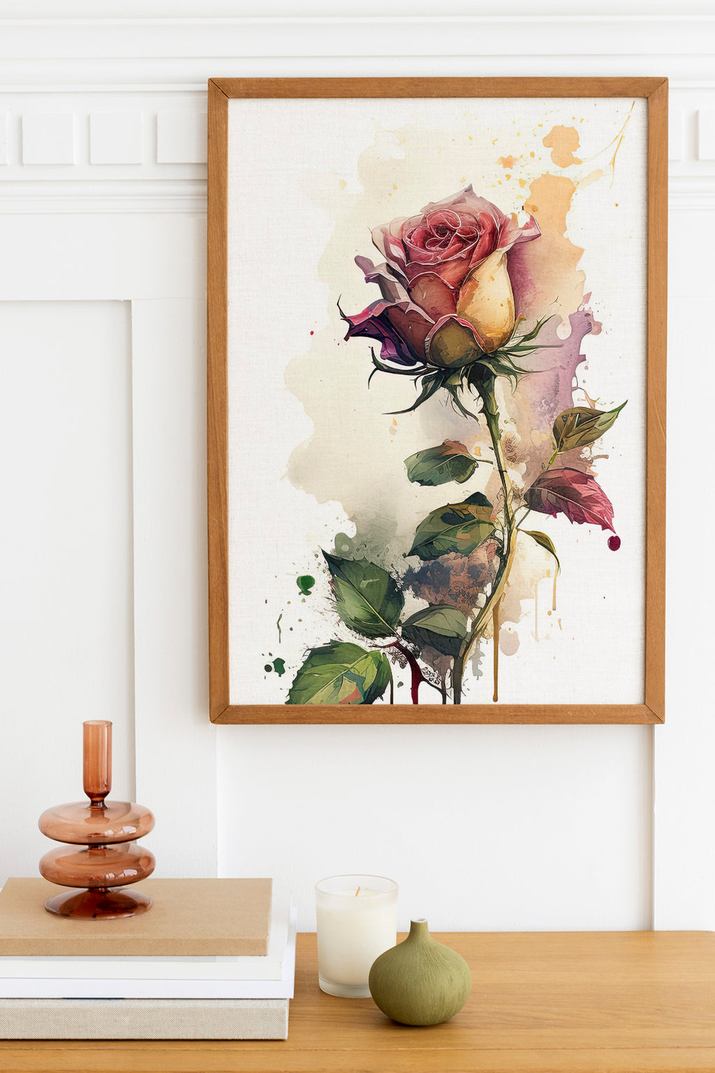 a framed painting of a vibrant pink rose with green leaves, hanging on a white wall above a wooden table. On the table, there is a stack of books, a small green vase, and a white candle.