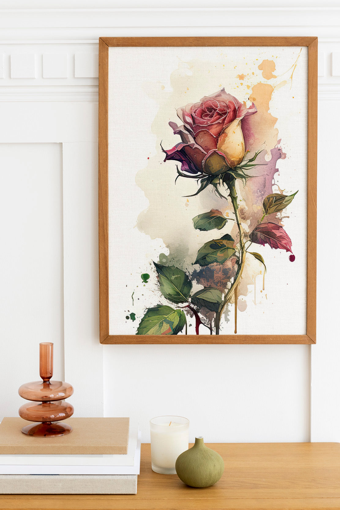 a framed painting of a vibrant pink rose with green leaves, hanging on a white wall above a wooden table. On the table, there is a stack of books, a small green vase, and a white candle.