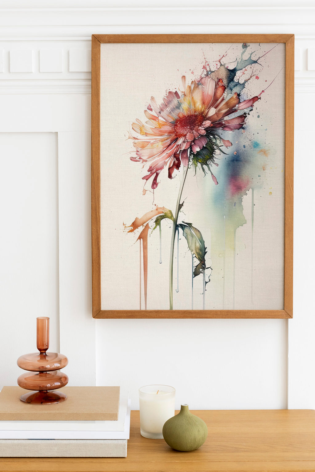 a framed watercolor painting of a vibrant pink and orange flower, hanging on a white wall above a wooden table. The table also holds a stack of books, a small green vase, and a white candle.