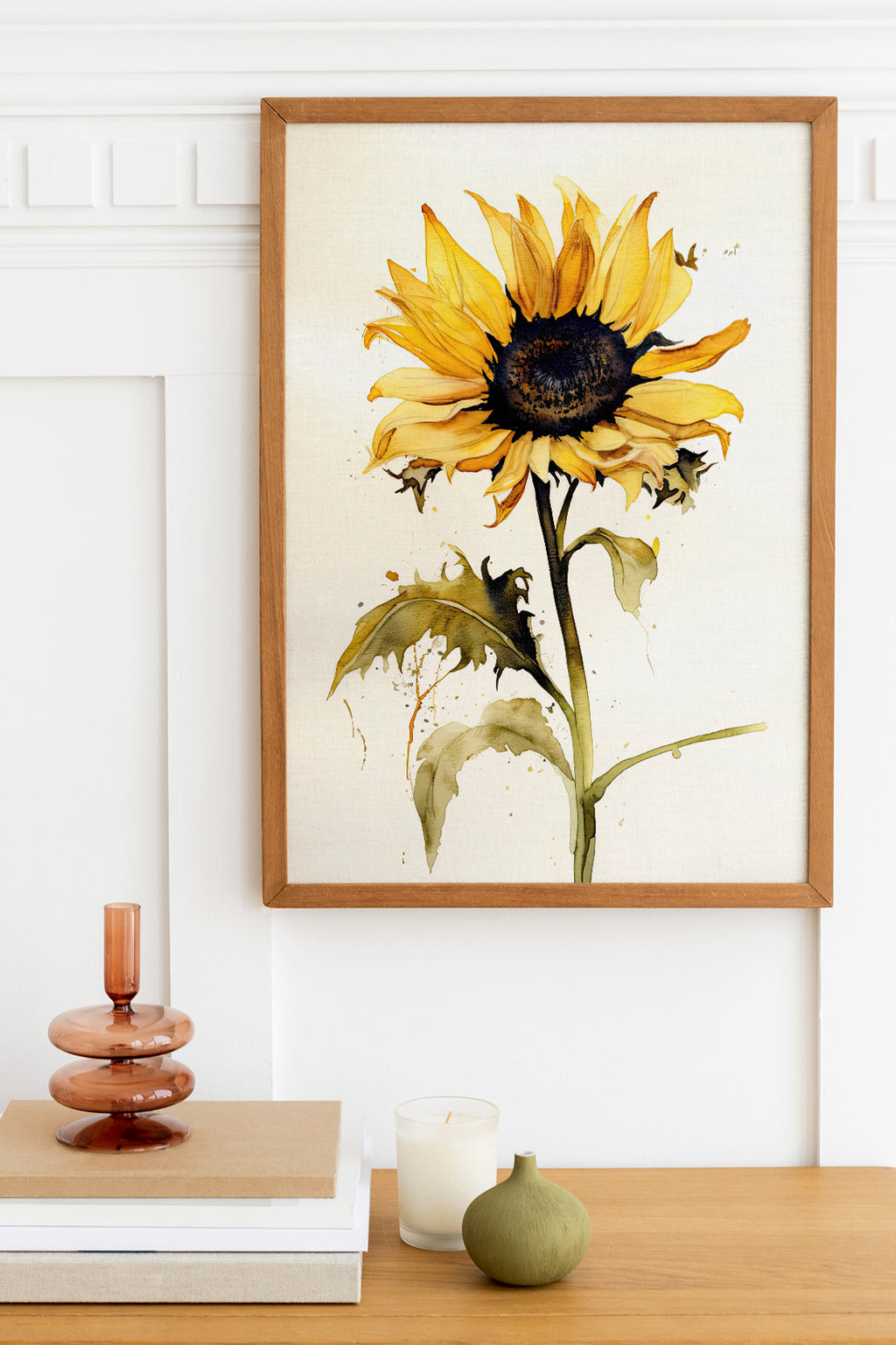 a framed painting of a yellow sunflower hanging on a white wall above a wooden table. On the table, there is a stack of books, a small green vase, and a white candle.
