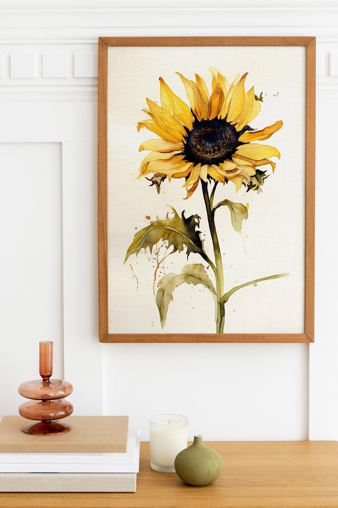 a framed painting of a yellow sunflower hanging on a white wall above a wooden table. On the table, there is a stack of books, a small green vase, and a white candle.