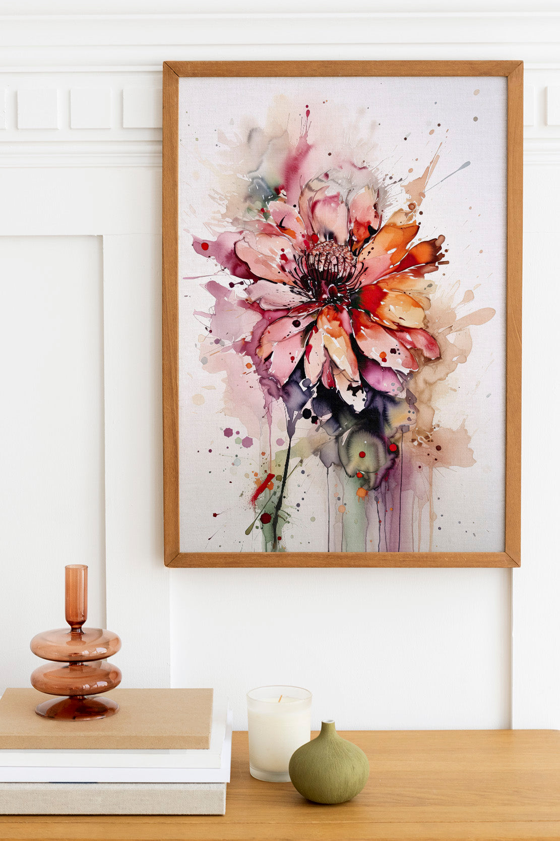 a framed watercolor painting of a vibrant pink and orange flower, hanging on a white wall above a wooden table. The table also holds a stack of books, a small green vase, and a white candle.