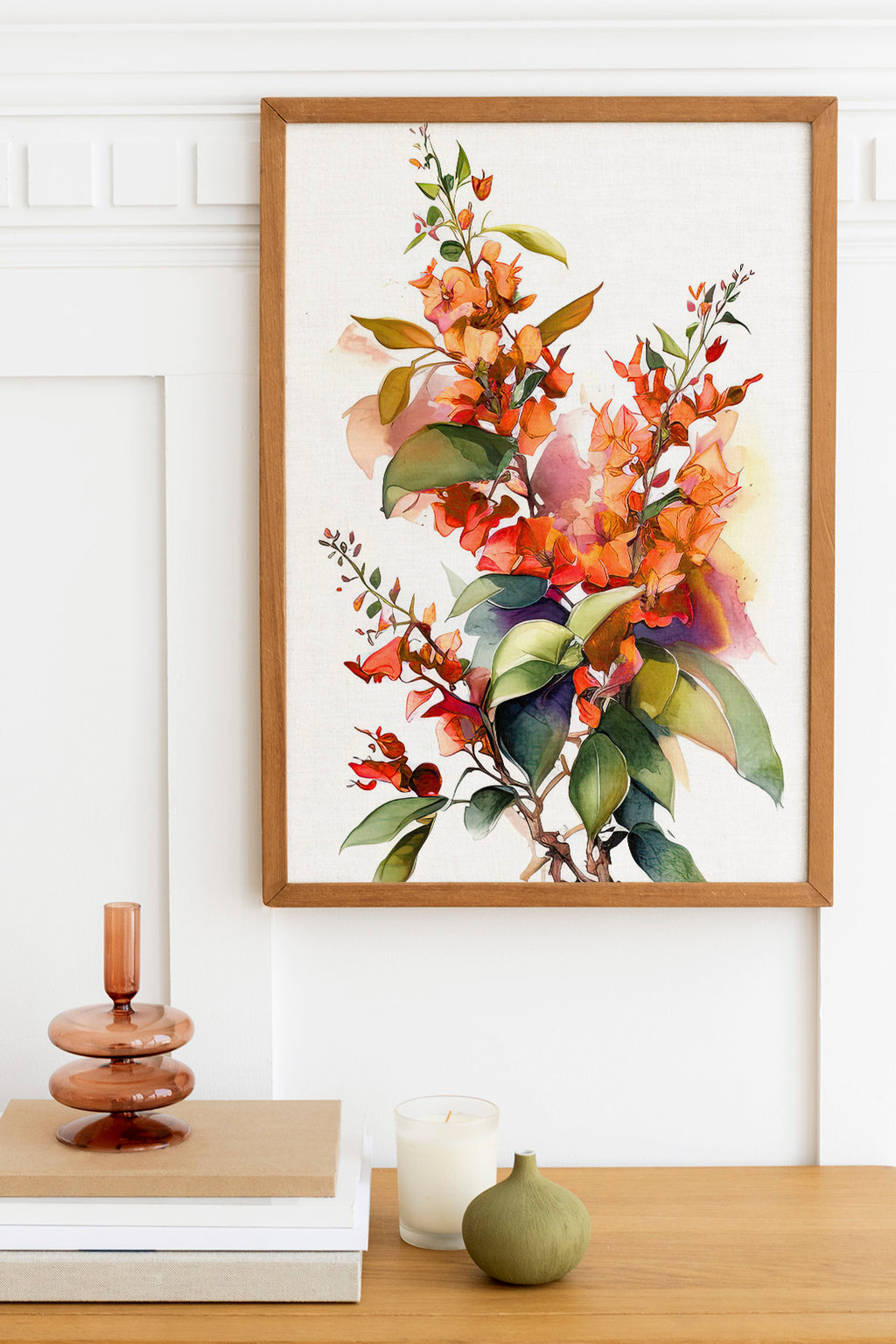 a framed painting of a vibrant floral arrangement hanging on a white wall above a wooden table. The table also holds a stack of books, a small green vase, and a white candle.