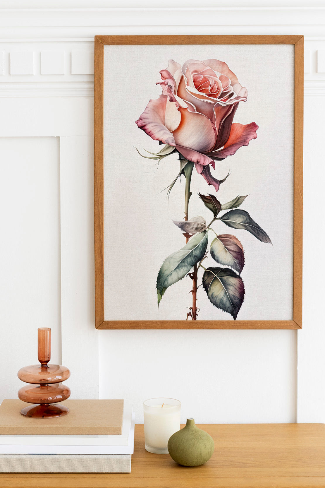 a framed painting of a pink rose hanging on a wall, with a wooden shelf below it holding a stack of books, a small green vase, and a white candle.