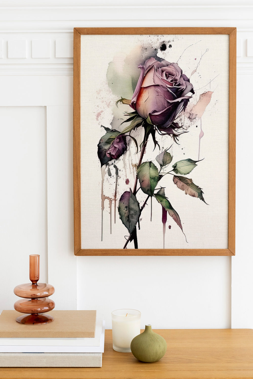 a framed painting of a purple rose with green leaves, hanging on a wall above a wooden table with various decorative items such as a candle, a small green vase, and a stack of books.