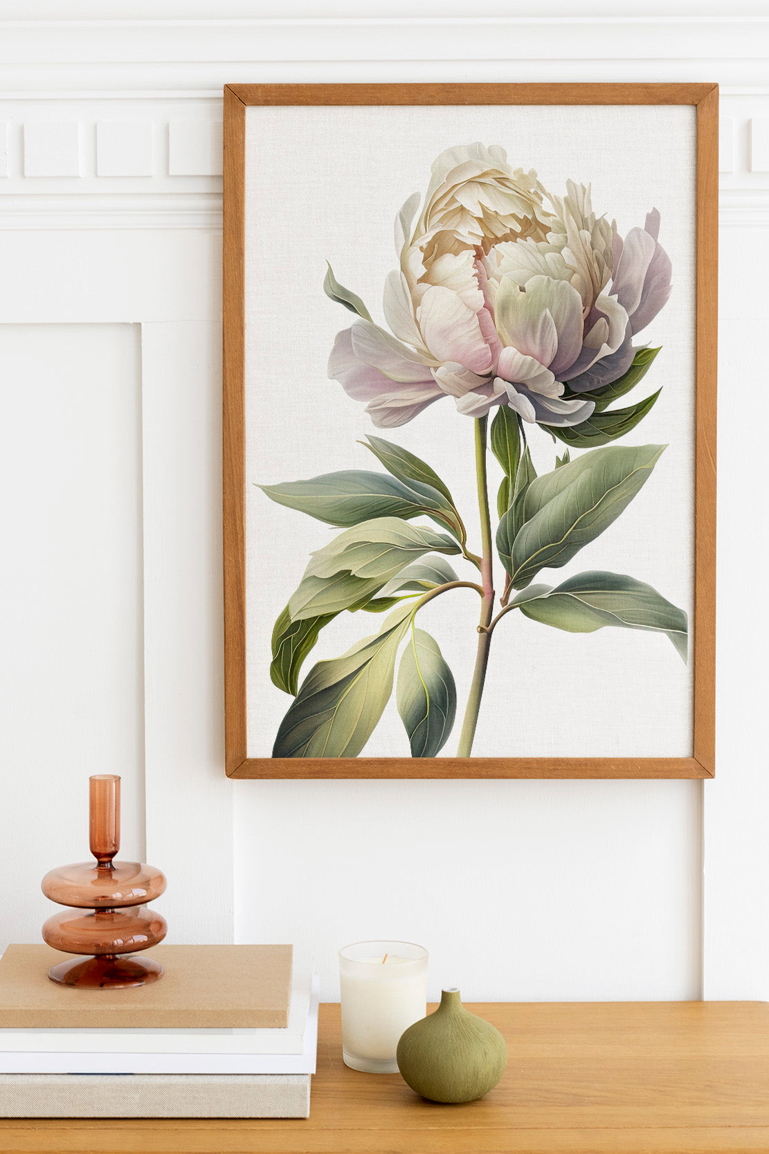 a framed painting of a large white peony flower hanging on a wall above a wooden table with various decorative items, including a candle, a small green vase, and a stack of books.