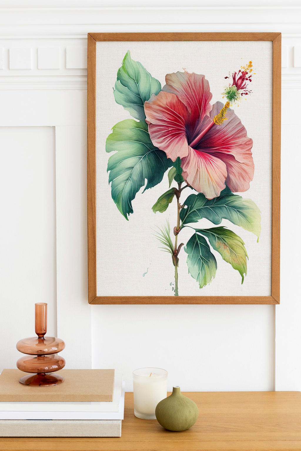 a framed painting of a large, vibrant hibiscus flower hanging on a white wall above a wooden table. The table also has a stack of books, a small green vase, and a white candle.