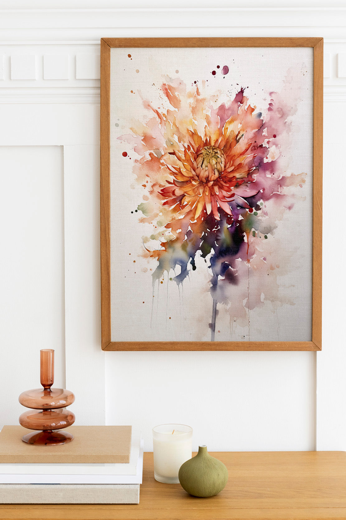 a wooden table with a framed watercolor painting of a vibrant orange and pink flower hanging above it. The table also has a stack of books, a small green vase, and a white candle.