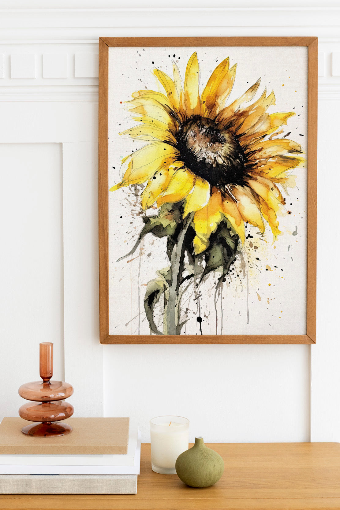 a framed watercolor painting of a vibrant yellow sunflower hanging on a white wall above a wooden table. The table also holds a stack of books, a small green vase, and a white candle.