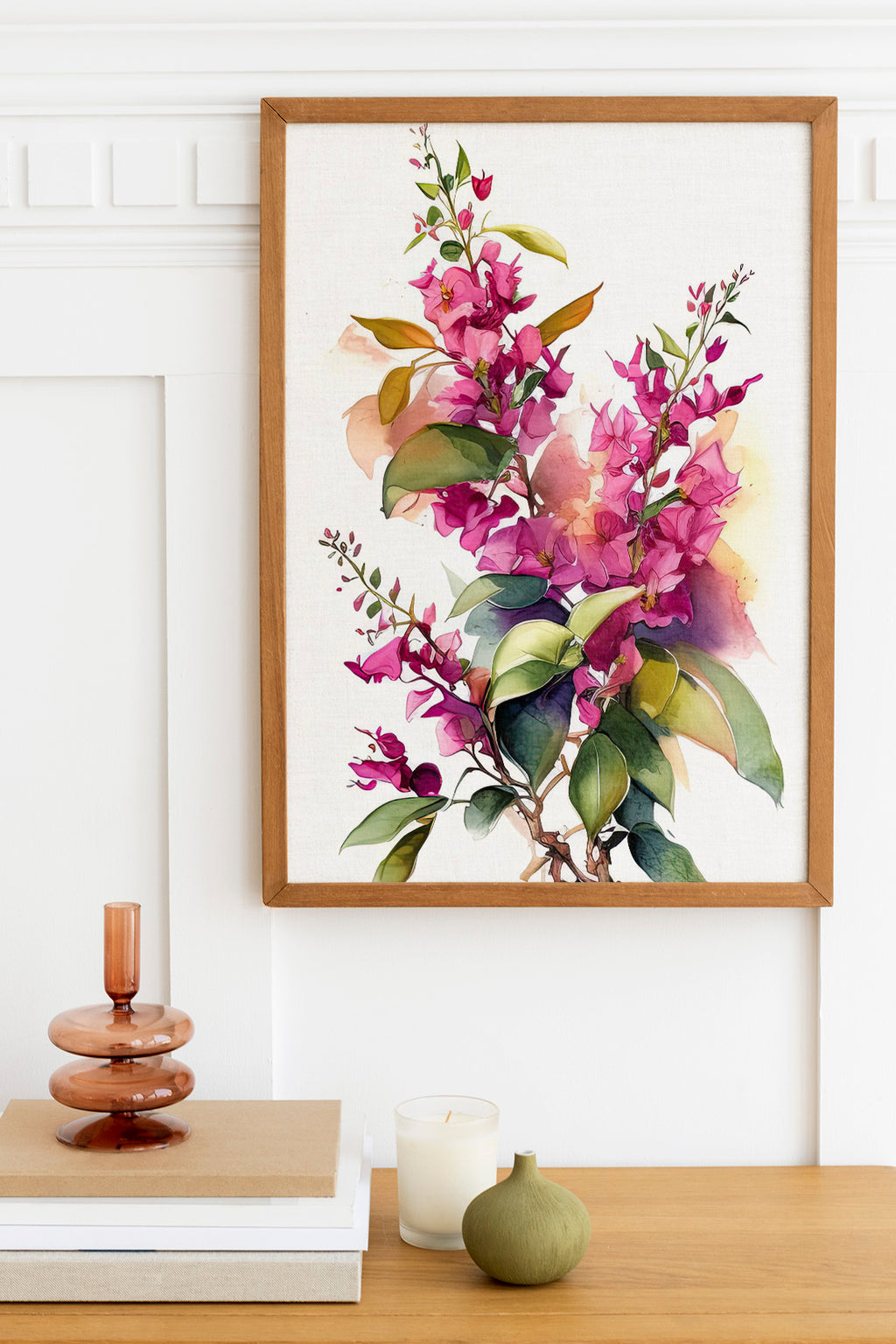 a framed watercolor painting of a branch with purple flowers and green leaves, hanging on a white wall above a wooden table. On the table, there is a stack of books, a small green vase, and a white candle.