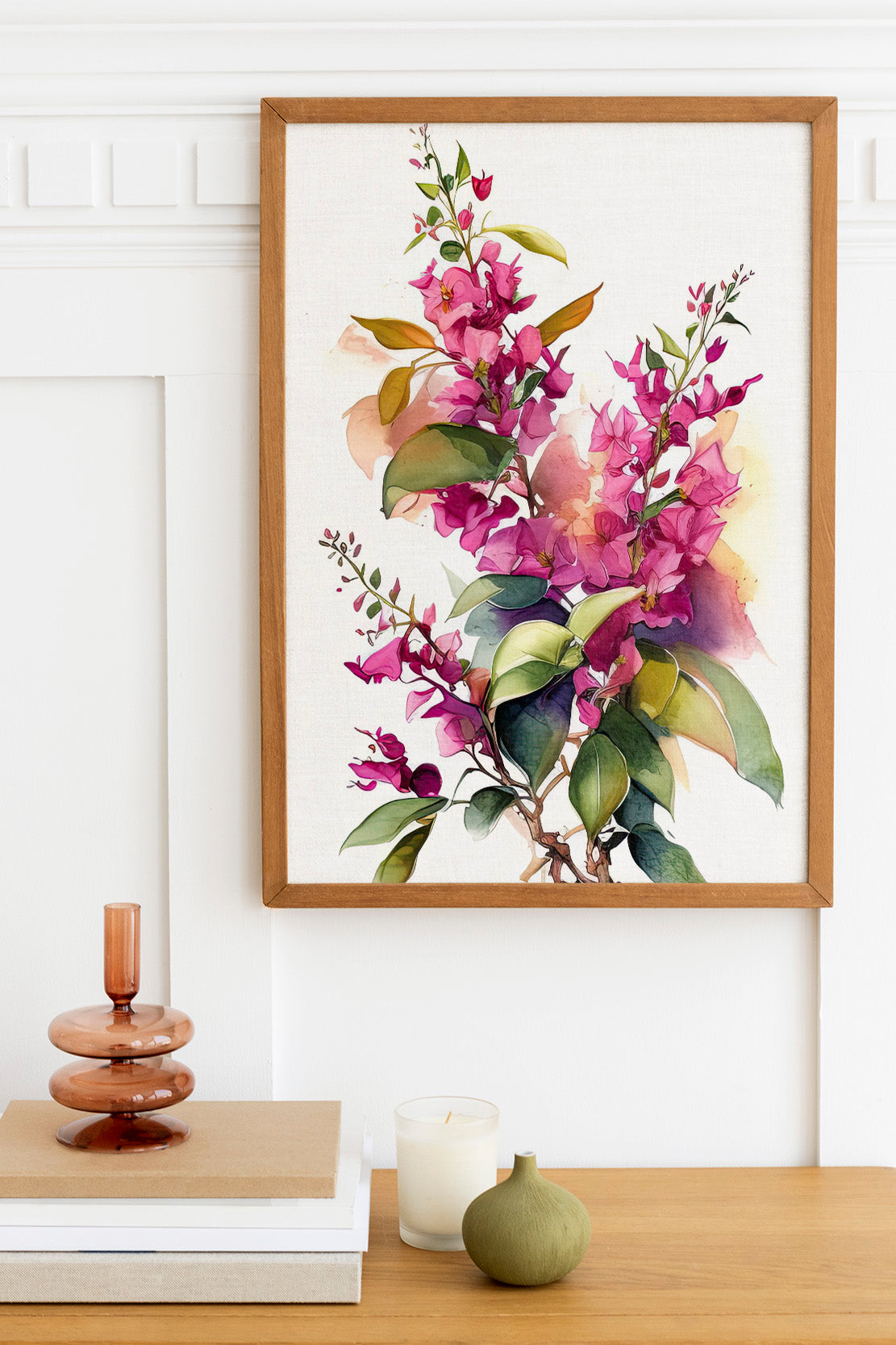 a framed watercolor painting of a branch with purple flowers and green leaves, hanging on a white wall above a wooden table. On the table, there is a stack of books, a small green vase, and a white candle.