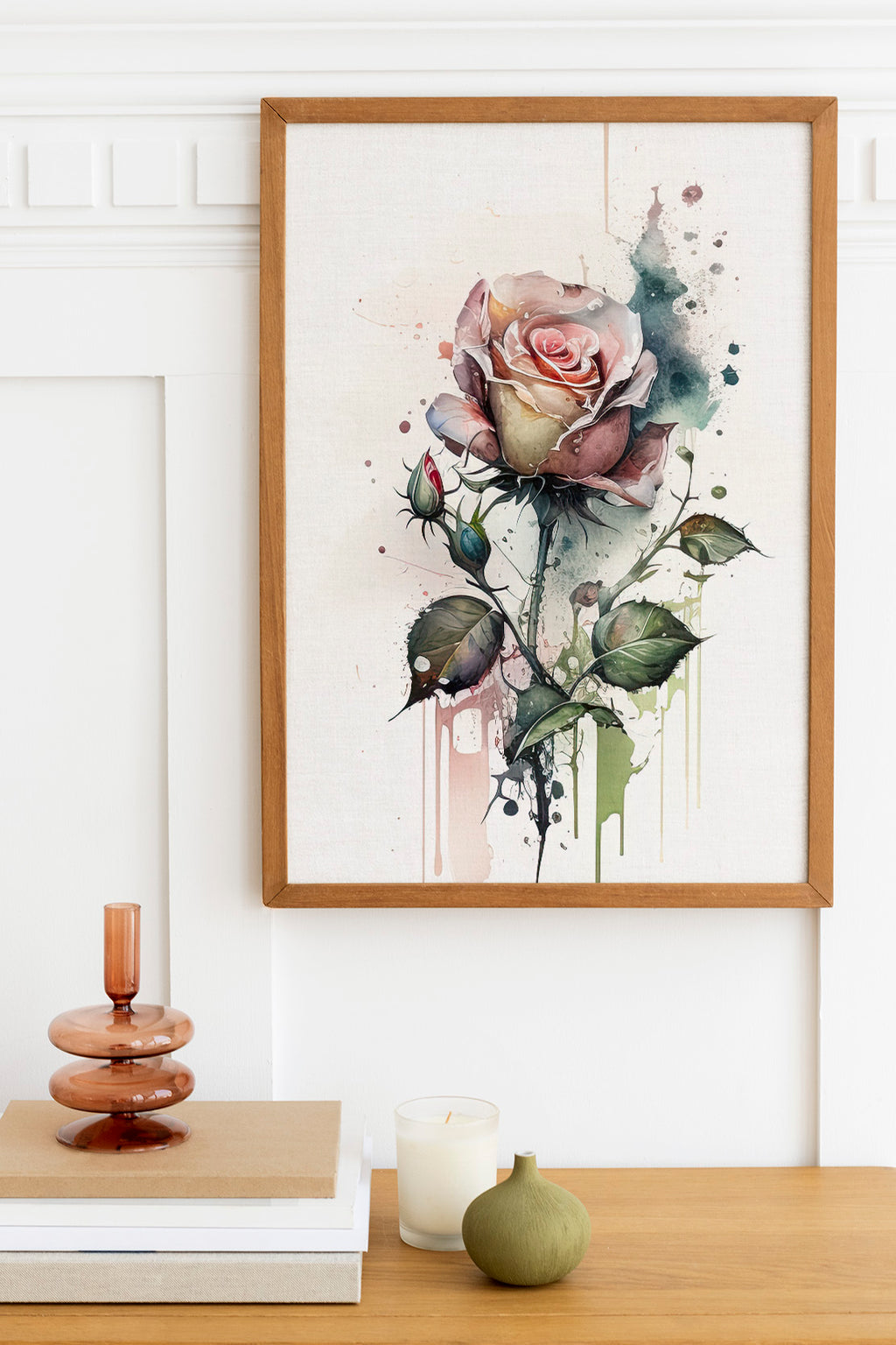 a framed watercolor painting of a pink rose with green leaves, hanging on a white wall above a wooden table with various decorative items, including a candle, a stack of books, and a small green vase.