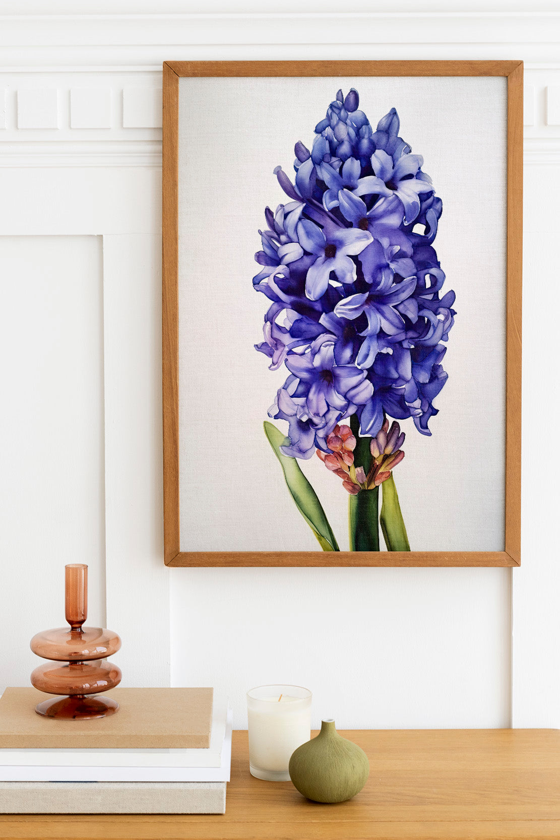 a framed painting of a large purple flower, which appears to be a hyacinth, hanging on a white wall above a wooden table. The table also has a stack of books, a small green vase, and a white candle.