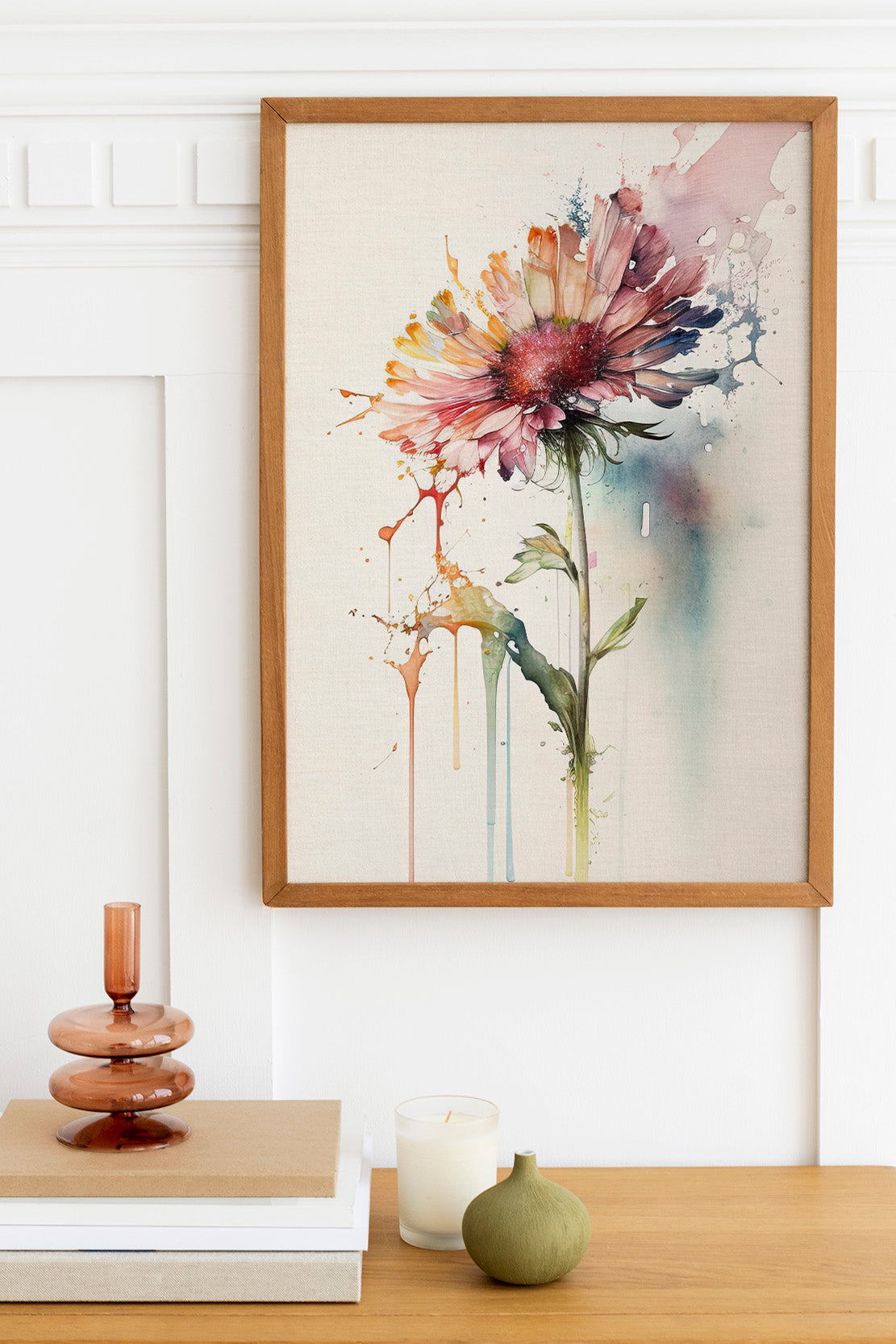 a framed watercolor painting of a flower hanging on a wall above a wooden table with various decorative items, including a candle, a stack of books, and a small green vase.