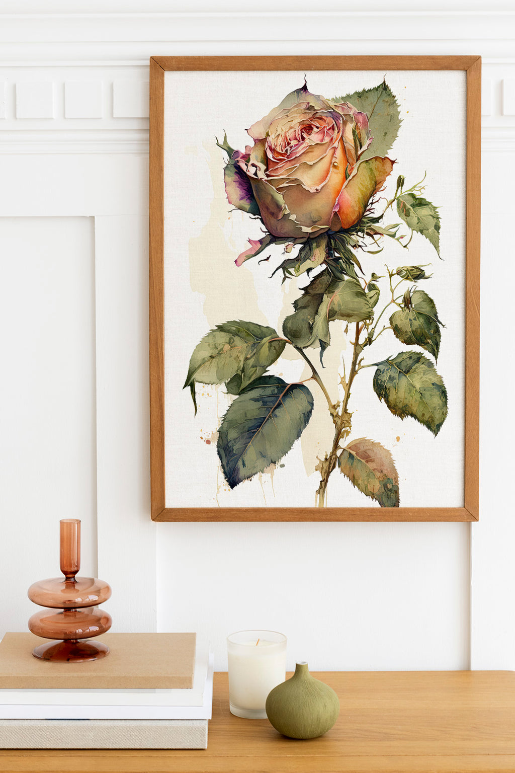 a framed painting of a large, vibrant rose with green leaves, hanging on a white wall above a wooden table. On the table, there is a stack of books, a small green vase, and a white candle.