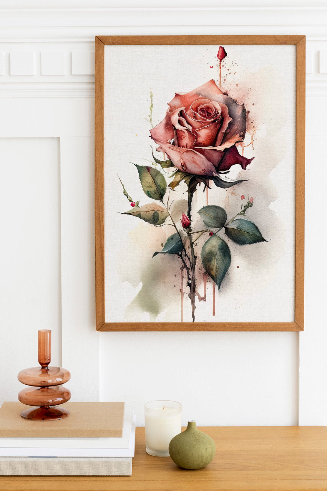 a framed painting of a red rose with green leaves and stems, hanging on a white wall above a wooden table with various objects on it, including a stack of books, a candle, and a small green vase.