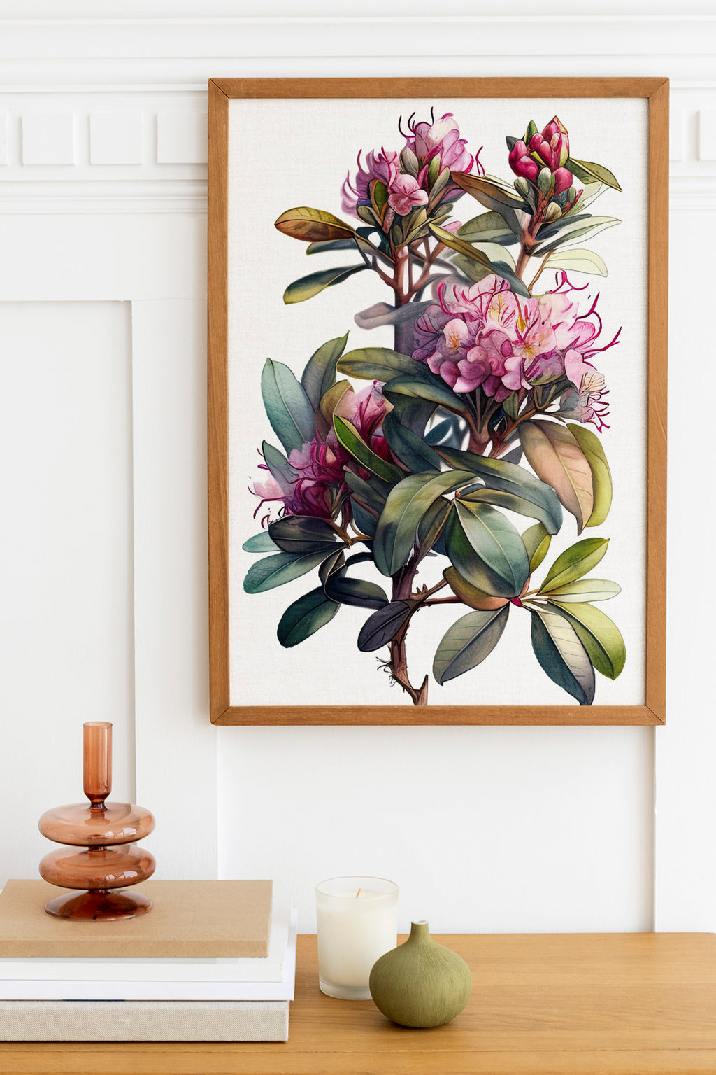 a framed painting of a vibrant floral arrangement hanging on a white wall above a wooden table. The painting showcases a variety of pink and purple flowers, along with lush green leaves. On the table, there is a stack of books, a small green vase, and a white candle, creating a cozy and inviting atmosphere.