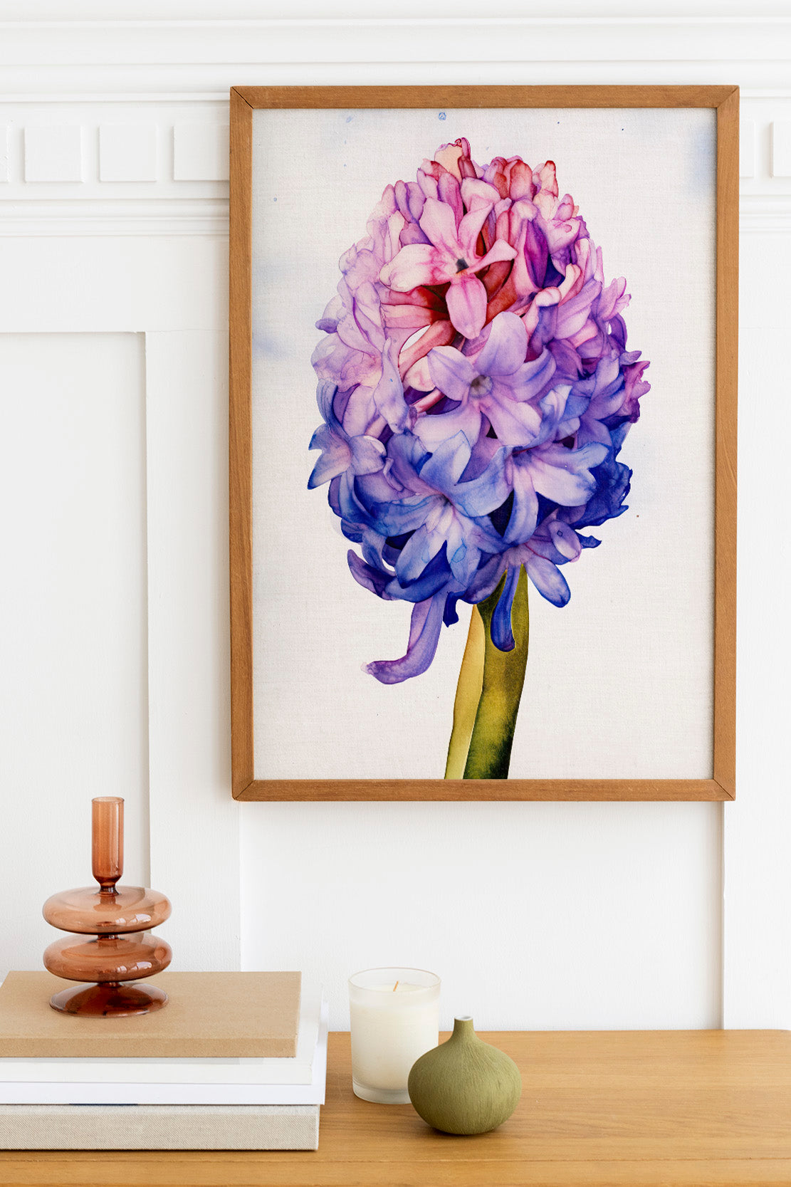 a framed watercolor painting of a large, vibrant purple flower with delicate petals, hanging on a white wall above a wooden table. The table also holds a small vase, a stack of books, and a candle.
