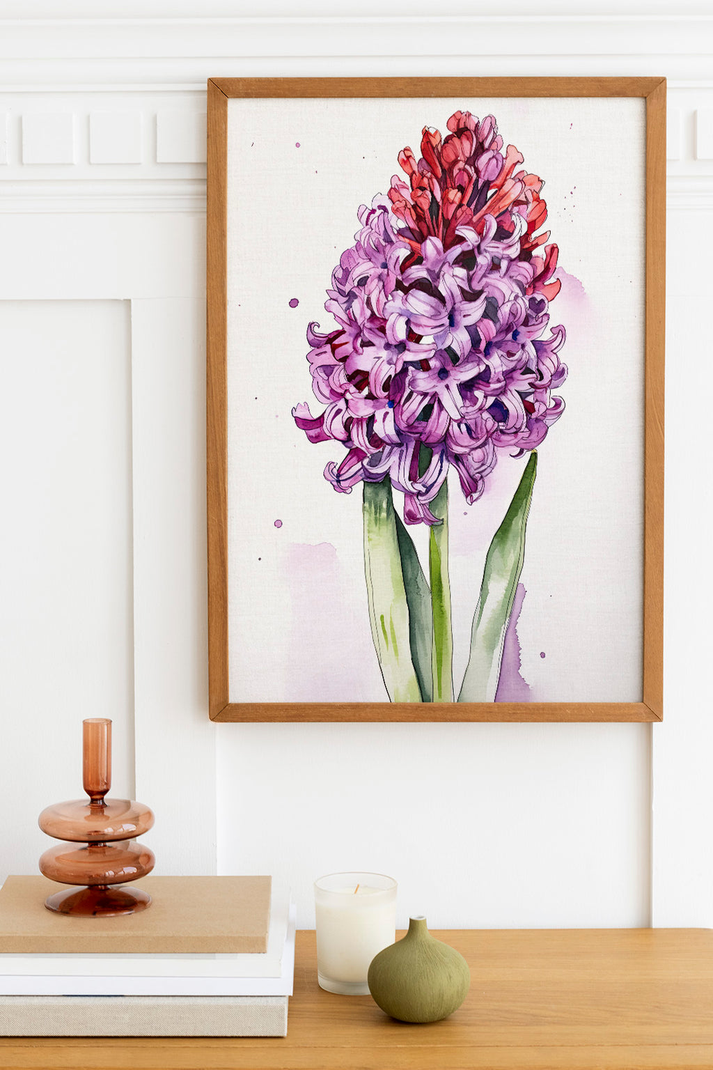 a framed watercolor painting of a large purple flower, hanging on a white wall above a wooden table. On the table, there is a stack of books, a small green vase, and a white candle.