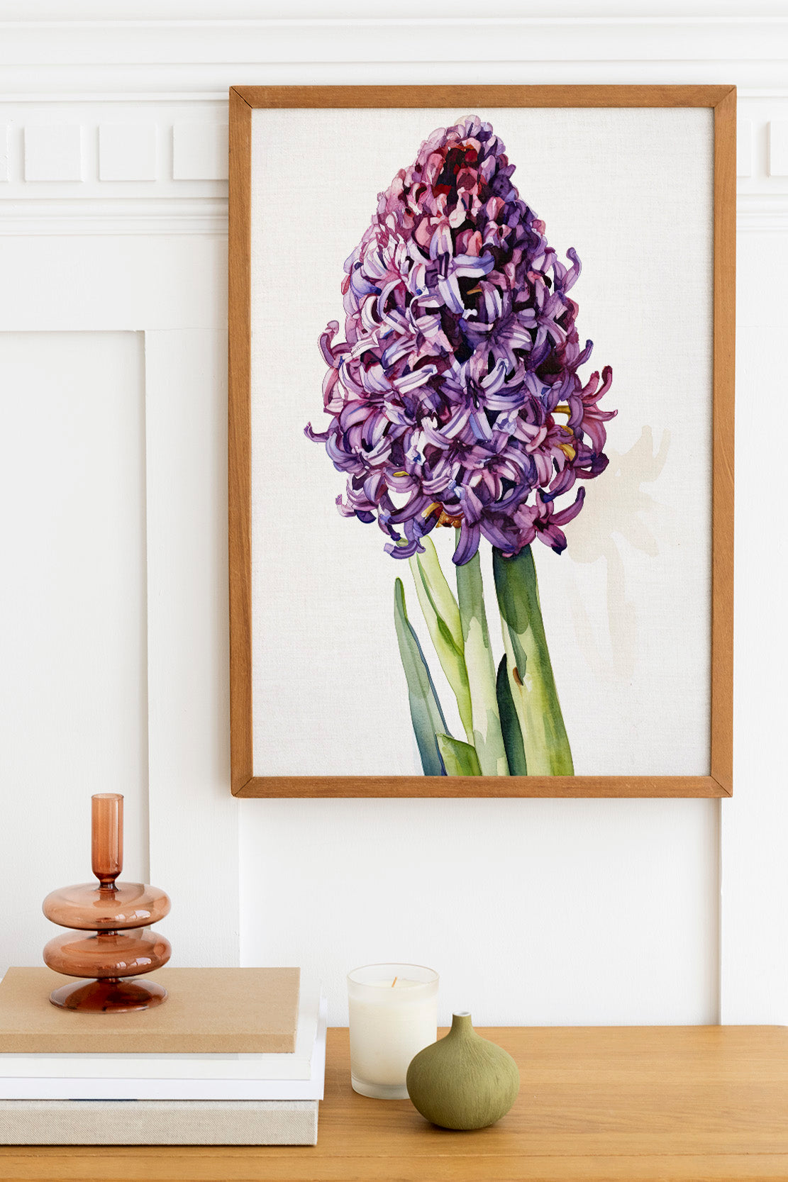 a wooden table with a framed painting of a purple hyacinth flower hanging above it. The table also has a stack of books, a small green vase, and a candle.