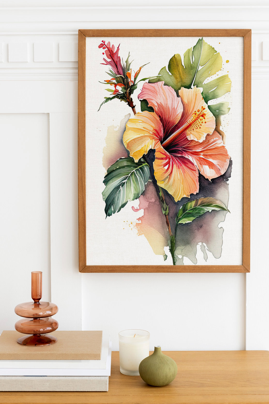 a framed watercolor painting of a vibrant hibiscus flower, with a wooden frame and a white background. The painting is hung on a white wall above a wooden table with various decorative items, including a stack of books, a candle, and a small green vase.