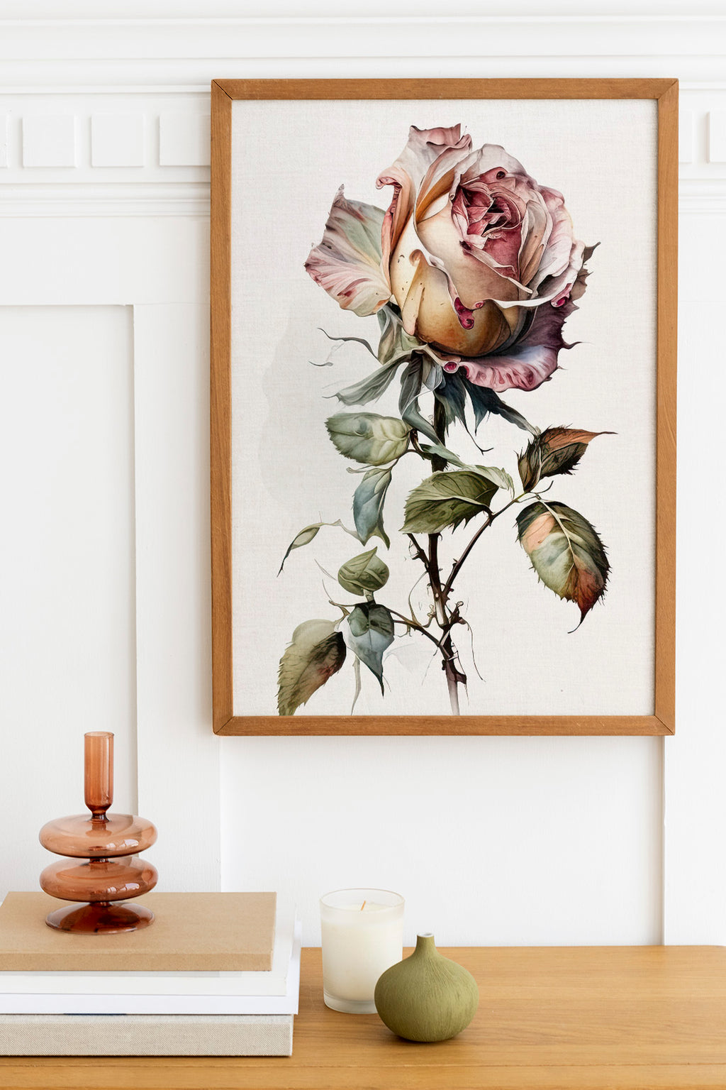 a framed painting of a large, vibrant rose on a white wall, with a wooden shelf below it holding a stack of books, a small green vase, and a white candle.