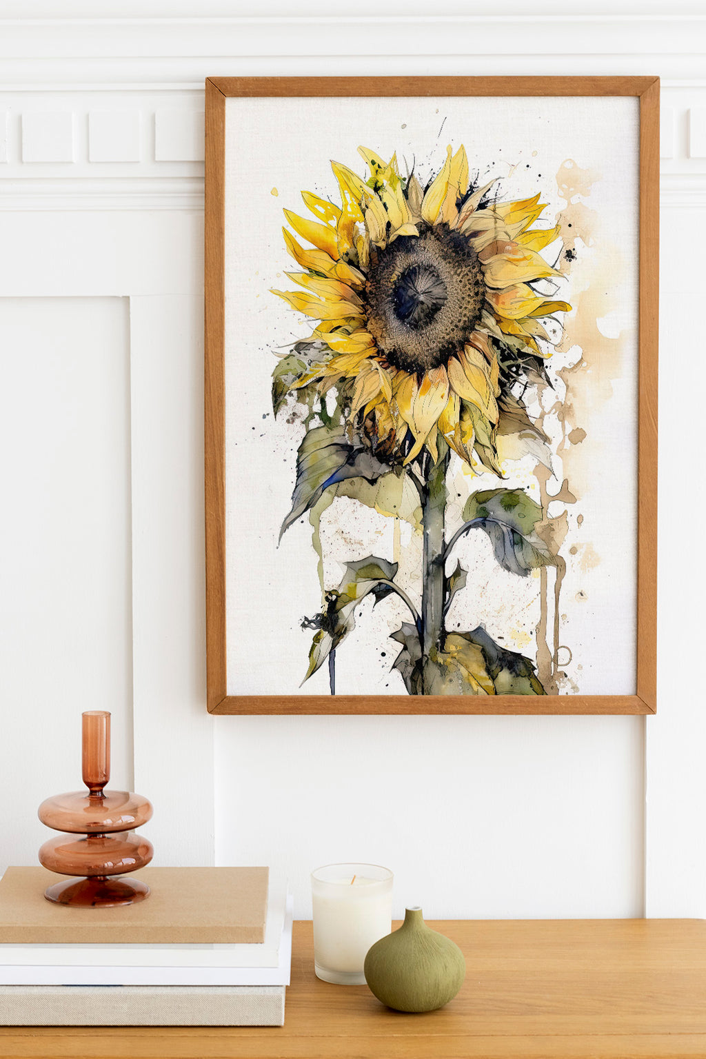 a framed watercolor painting of a sunflower hanging on a wall above a wooden table with various decorative items, including a candle, a stack of books, and a small green vase.